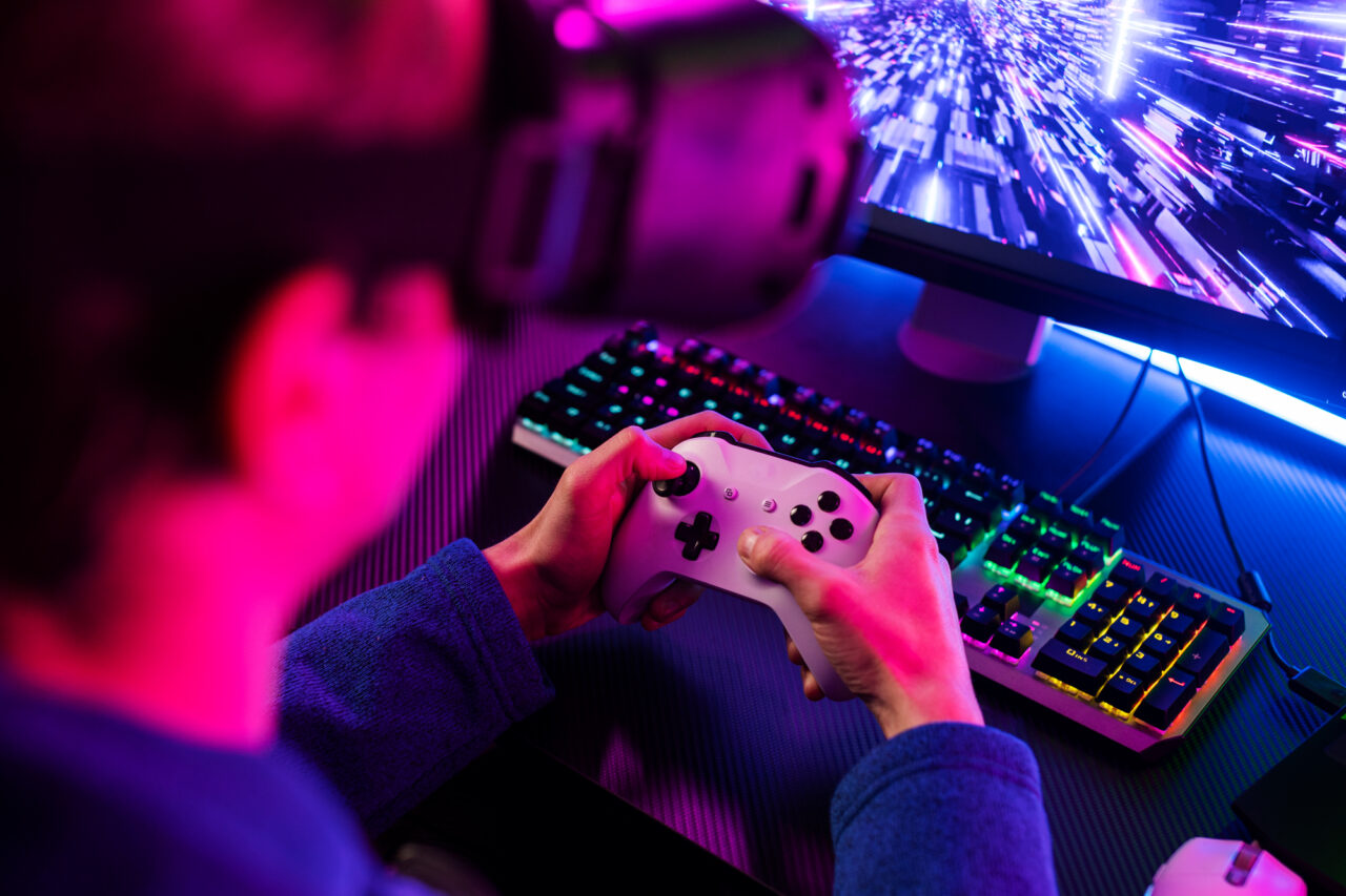 Person with VR headset holding a game controller, neon-lit gaming setup with colorful keyboard and monitor